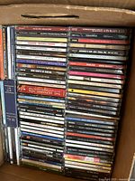 Top view of a box filled with over 70 music CDs mostly pop and rock genres, stacked horizontally revealing titles and artist names on spines.