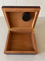 Open wooden cigar humidor showing interior with attached round humidifier on inside lid and brass hinges.