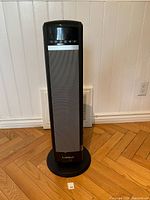 Full front view of black Lasko tower heater on wooden floor against wall with control panel visible at top.