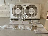 Front view of NOMA dual window fan showing two fan blades, control dials, power cord, accessories and manuals on countertop.
