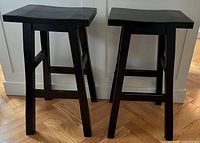 Two matching black wooden counter stools placed side by side on parquet flooring against a white paneled wall, showing overall design and condition.