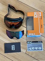 Goggles lying flat showing amber installed lens, swappable reflective blue-green lens, original orange Spy Ace box, instruction manual and installation instructions.