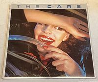 Front cover of The Cars vintage vinyl album showing woman with hand shielding eyes and bright red lipstick and nails holding steering wheel.