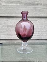 Front view of an amethyst colored glass vase with a narrow top and clear glass twisted stem and base, placed against a horizontal wood panel background.