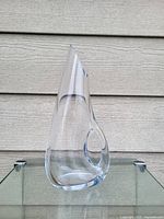 Front side view of the clear pierced crystal glass decanter with a circular handle near the top and a tapered spout, resting on a glass surface.