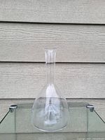 Clear glass decanter with narrow neck and bulbous base shown against a gray wall and glass table.