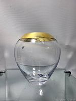 Front view of clear glass vase with smooth rounded body and wide gold band trim at top rim, placed on glass shelf with white background.