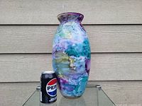 Front angled view of large, colorful glass flower vase next to a Pepsi can for size comparison on a glass surface against a horizontal siding background.