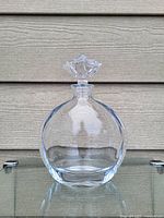 Clear glass bottle with flower-shaped stopper upright on a glass shelf in front of horizontal siding wall.