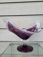 Large purple glass pedestal display with clear glass accents on a glass surface in front of beige siding.