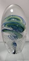Front view of oval glass paper weight showing green and blue layered swirl design with bubbles inside.