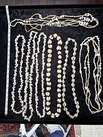 Photo of 11 natural seashell beaded necklaces laid flat on black fabric background with measurement tape showing lengths.