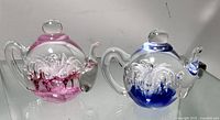 Photo showing two glass paperweights, one shaped like a teapot with pink interior glass detail and another round one with a swan inside and blue glass detail.