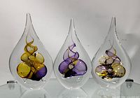 Front-facing view of three teardrop shaped glass paper weights with abstract designs inside in yellow, purple, black and gold colors on clear glass bases.