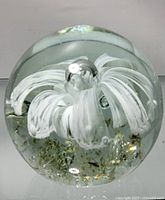 Top and angle views of the glass paper weight showing the white flower design inside with bubbles and gold flecks.