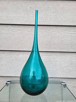 Front view of turquoise blown glass bulb vase with long, thin neck standing against light brown siding.