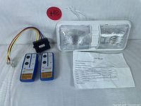Photo showing the LED light fixture with two clear lens panels, two remote controls, wiring harness, and user manual arranged side by side.