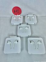 Five individually sealed, white wired earbud sets arranged on a white surface, each in its own white plastic packaging.