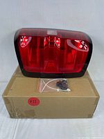 Front view of the brand new red driver side tail light assembly resting on a cardboard box with mounting hardware included in a plastic bag.