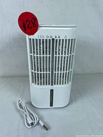 Front view of white portable air conditioner with USB cable coiled beside it. Shows vertical air vents, water tank window, and control icons on top.