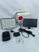 Lot contents laid out on striped white surface: rectangular LED light panel with 294 LEDs and motion sensor, black solar panel connected by cable, black charging base, small mounting bracket, and a pack of screws and anchors, with original packaging box visible in one photo.