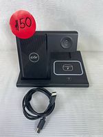 Front view of black EXW 3-in-1 wireless charging station with three charging areas: vertical phone stand, circular watch charger, flat earbud pad. USB-C cable placed in front.