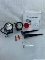 Two black LED landscape spotlights, two black plastic ground stakes, three orange wire connectors, two metal hex keys, and the original white box labeled 'LED Landscape Lighting'. Pieces arranged on white surface.