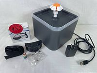 Photo of the automatic pet water fountain with flower-shaped spout, power adapter, USB cable, no-bark collar strap, transmitter, batteries, and packaging box with instructions.