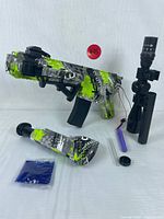 Electric gel ball blaster in green, black, white camo pattern with accessories laid out including scope, battery, gel balls, and detachable stock.