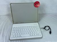 White Bluetooth keyboard attached to light grey folio tablet case, laid open revealing keyboard layout and charging cable nearby.