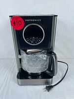 Full view of TaoTronics coffee maker with bubble wrap inside the glass carafe and power cord attached, showing front control panel with buttons.