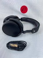 Photo showing the black wireless over-ear headphones lying flat and a black charging cable placed in front of them on a white surface.