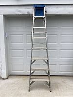 Full height image of 8 foot aluminum step ladder standing open against garage doors, showing metal steps and blue plastic top tray.