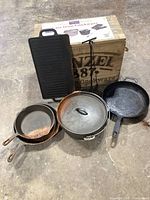 Photo showing full cast iron cookware set including two skillets, covered pot, griddle pan, and frying pan placed on floor with wooden box in background