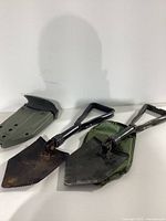 Two folding metal entrenching shovels with serrated edges and foldable handles, shown with olive green plastic sheath.