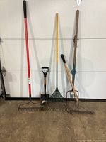 Full view of five garden tools: metal rake with red handle, shovel with D-grip, leaf rake with yellow handle, garden hoe, and weed cutter against a white door.