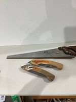 Craftsman hand saw with wooden handle and two folding pruning saws with wooden handles laid on a white surface.