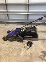 Side view of the Kobalt cordless lawn mower with grass bag attached and charger placed in front on the floor.