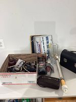 Image showing various items including a USPS box containing assorted tools, metal pipe tool, clear glass cylinder vase, black leather case, and some collectible model cars and accessories.