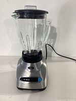 Full view of the Oster blender on a flat surface, showing the glass jar, black lid, silver base, and power cord.