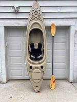 Full view of tan Pelican kayak propped against garage wall with included paddle upright beside it