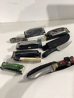 View of various folding pocket knives and one fixed blade with sheath, displayed on white surface showing brands and boxes.
