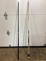 Two full-length pool cues leaning against a door, one black with wrapped grip and one natural wood finish