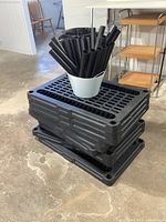 Stack of 10 black plastic shelving shelves with grid surface and a white container holding hollow black plastic legs on top.