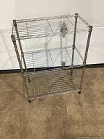 Front view of the three-tier wire metal shelving unit on a concrete floor against a white wall showing the chrome finish and open wire shelving design.