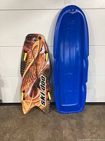 Two snow sleds standing upright; one is a Ski-Doo branded sled with colorful graphics and yellow hand grips, the other is a plain blue plastic sled with molded shape.