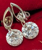 Close-up photo showing one earring with a teardrop-shaped 10KT gold mounting holding a clear round faceted gemstone, against a red background.