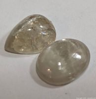 Two genuine color change Zultanite gemstones on a white surface, one pear shaped and one oval cabochon, showing internal features and polish.