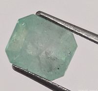 Close-up of the loose rectangular step-cut emerald held by tweezers showing light green color and internal inclusions