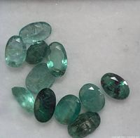 Close-up photo of 11 oval cut green emerald gemstones with faceting, some showing natural inclusions and imperfections, arranged on a white background.